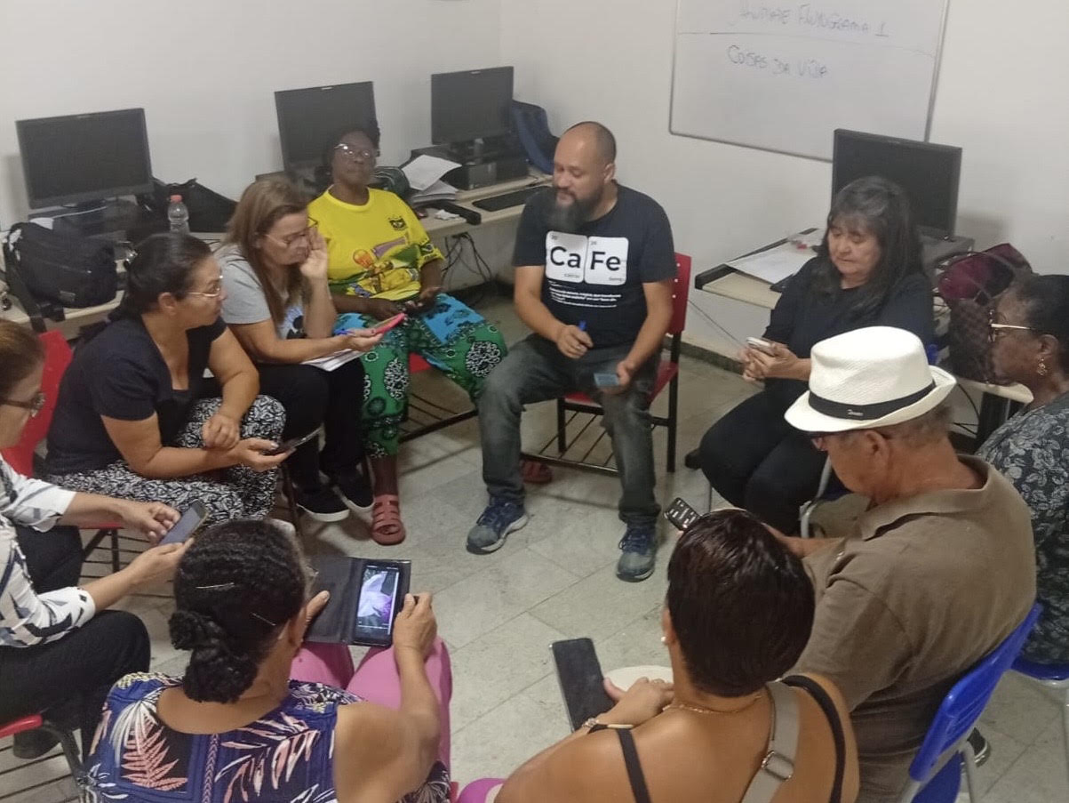 Em uma sala um grupo de pessoas sentam em cadeiras em roda, algumas conversando entre si e outras usando o celular. Atrás das pessoas existem computadores em mesas. 