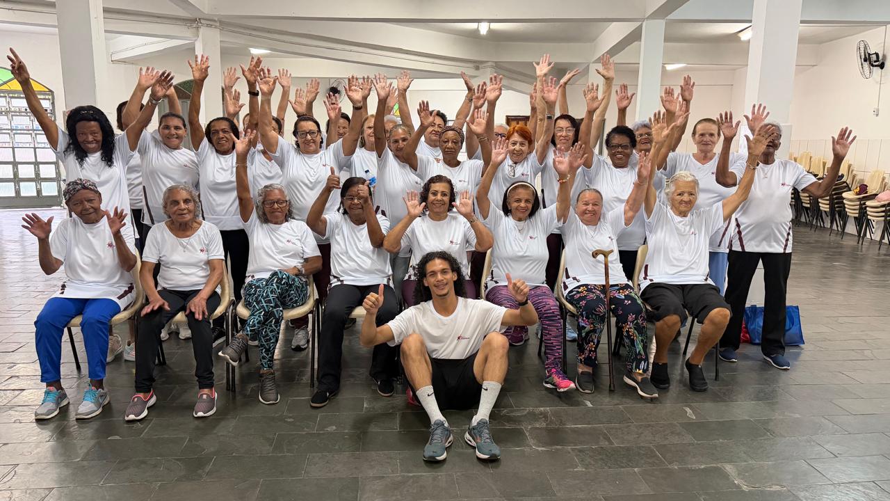 Vida Ativa transforma a rotina de idosos em Belo Horizonte com exercícios e inclusão Participantes do Programa Vida Ativa, da Prefeitura de Belo Horizonte, reunidas em um salão comunitário. Vestindo camisetas brancas do programa, as mulheres aparecem sorrindo e com os braços levantados, ao lado de um professor de Educação Física sentado à frente do grupo. A imagem registra um momento de integração, alegria e convivência entre as participantes.