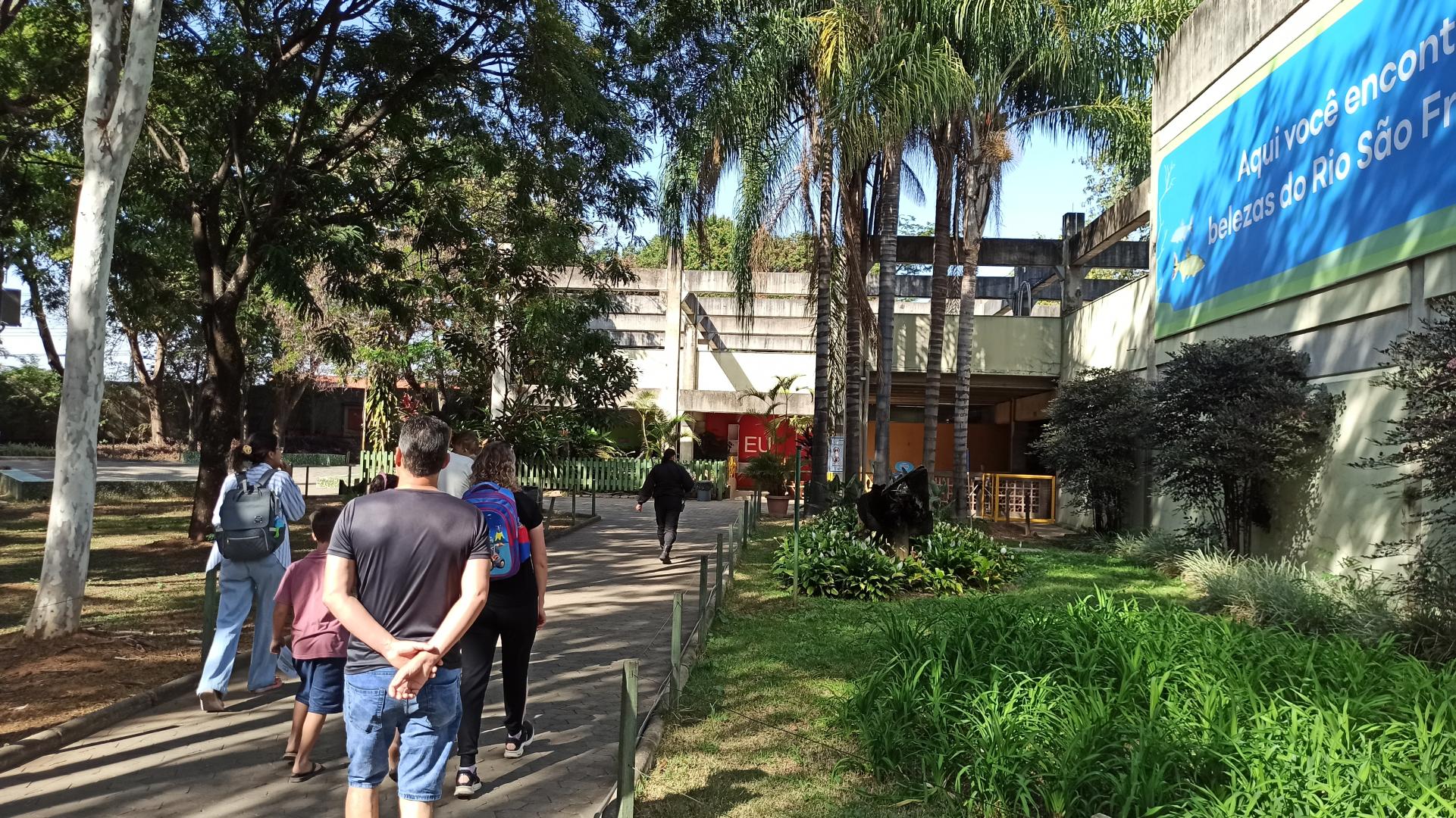 Aquário do Rio São Francisco promove atividade interativa sobre fenômeno da migração de peixes Visitantes caminham em direção ao Aquário do Rio São Francisco, no Zoológico de BH, por um caminho arborizado e cercado por jardins. À direita, um painel azul com ilustrações de peixes destaca as belezas do rio.