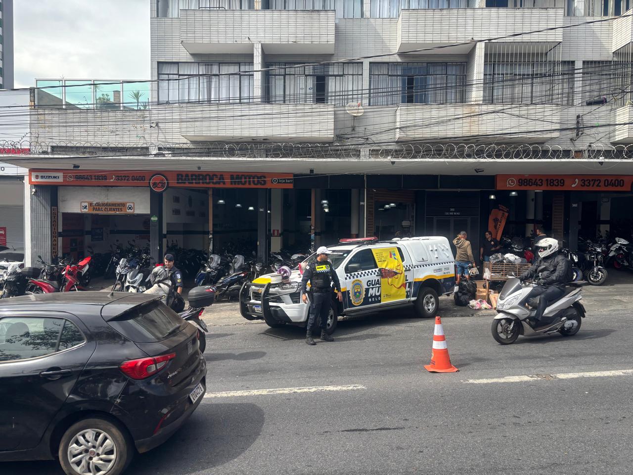 Guarda Municipal encerra Semana Nacional do Trânsito com blitz educativa para motociclistas Uma viatura da Guarda Municipal de Belo Horizonte encontra-se estacionada em frente a um estabelecimento comercial, com agentes ao redor e materiais da blitz educativa posicionados na viatura. A circulação do trânsito no local é orientada por cones de sinalização, enquanto carros e motocicletas transitam ao redor.