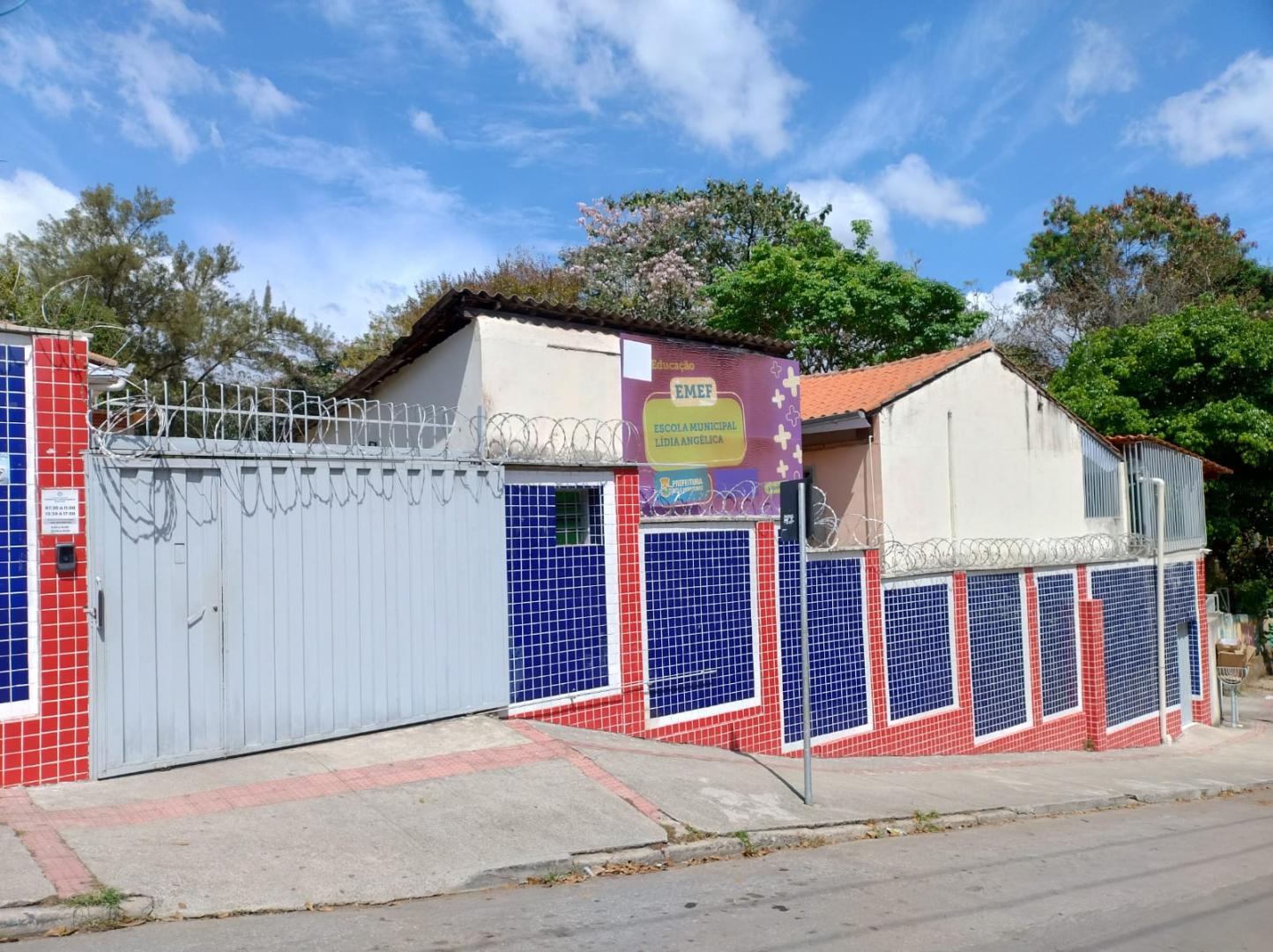 Escola Municipal Lídia Angélica vai passar por reforma para ampliação e modernização Fachada da Escola Municipal Lídia Angélica. O imóvel é térreo, com paredes revestidas por pastilhas nas cores azul, vermelho e branco e portões metálicos. A entrada e os muros são protegidos por concertina. Na fachada há uma placa de identificação e a logomarca da Prefeitura. O prédio possui telhados de diferentes alturas, janelas com grades e uma área de acesso com rampa de concreto. Em frente à escola, há uma calçada e a rua. Ao fundo, tem árvores de grande porte sob. céu azul com nuvens