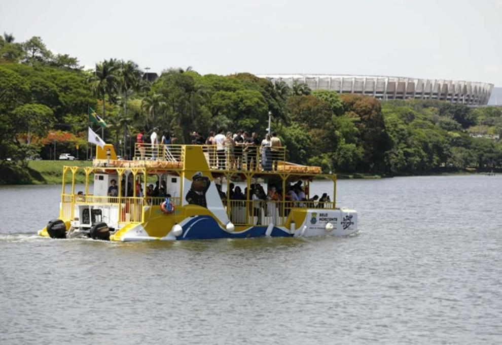 Foto de um barco com tonalidade amarela, azul e branca tansportando pessoas em navegação por espelho d'água, com o Mineirão ao fundo