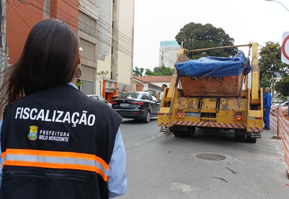 Fiscalização combate instalação irregular de caçambas com mais de 1,7 mil vistorias neste ano em BH Agente de fiscalização da Prefeitura de Belo Horizonte, de costas, usando colete de trabalho na cor azul, com listra alaranjada, acompanha caminhão com caçamba em uma rua com área de obras isolada.