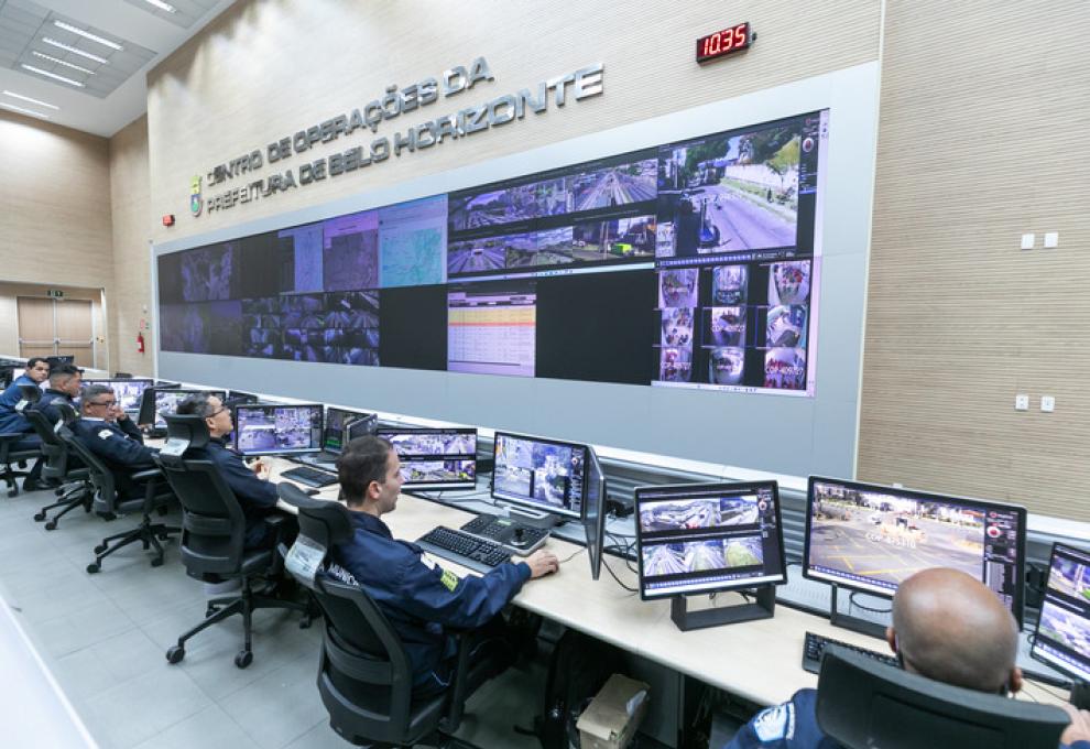Cinco agentes da Guarda Municipal de BH sentados em frente a monitores de computador na mesa e um painel maior na parede que mostram imagens de diferentes locais de Belo Horizonte 