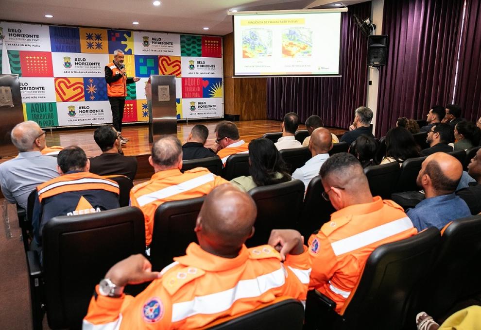 Imagem mostra várias pessoas uniformizadas com coletes laranja assistindo a uma apresentação de um homem tambémn de colete laranja. No fundo há um banner com logomarcas da PBH.