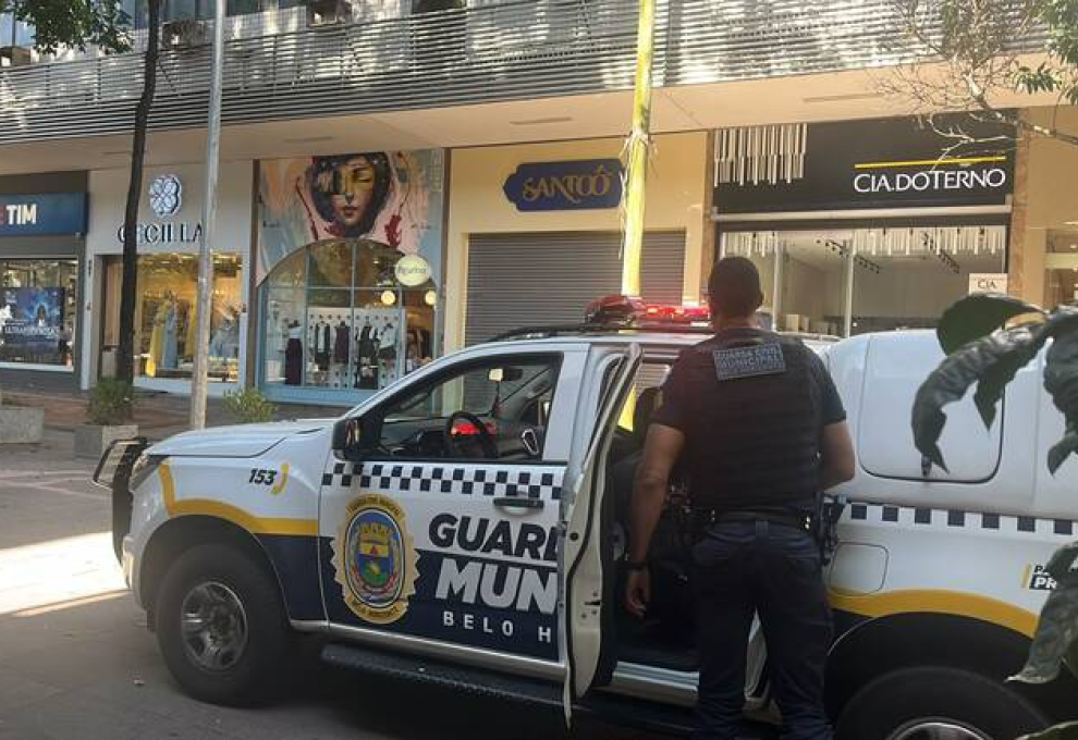 Viatura da Guarda Municipal de frente para três lojas, na rua. Agende da Guarda, uniformizado, parado ao lado da viatura, que está com a porta traseira aberta.
