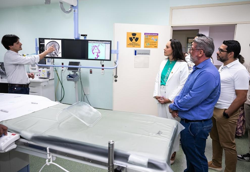 Hospital Célio de Castro amplia tratamento de AVC e passa a oferecer trombectomia mecânica pela rede pública de saúde de BH Imagem mostra uma maca em primeiro plano e atrás dela um homem apontando para duas telas de um aparelho do Serviço de Trombectomia Mecânica. O secretário de Saúde, de camisa azul, obserba, ao lado de uma mulher de camisa verde e branca. Há um homem de camiseta branca.