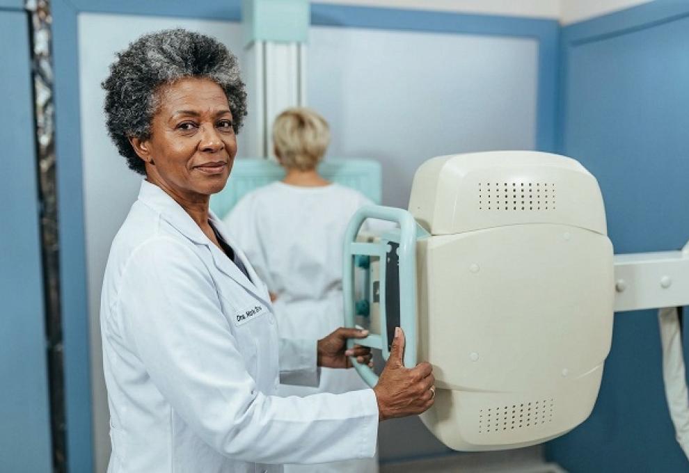 Foto de uma sala de exames de saúde. Em primeiro plano, uma profissional de saúde negra, de cabelos grisalhos e curtos, veste um jaleco branco e segura o equipamento de mamografia. À direita, aparece o aparelho de mamografia, de cor branca, com uma estrutura vertical e uma área de apoio para posicionamento da paciente. Ao fundo, uma mulher, de cabelos claros e usando roupa branca, está de costas diante do equipamento. 