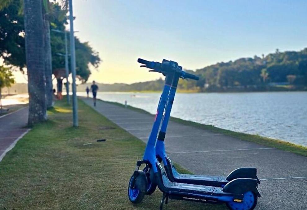 Patinetes elétricos chegam à Pampulha neste domingo (19) Patinete elétrico na cor azul, ao fundo, Lagoa da Pampulha e pôr-do-sol, enquanto pessoas correm e caminham na Orla da Lagoa da Pampulha