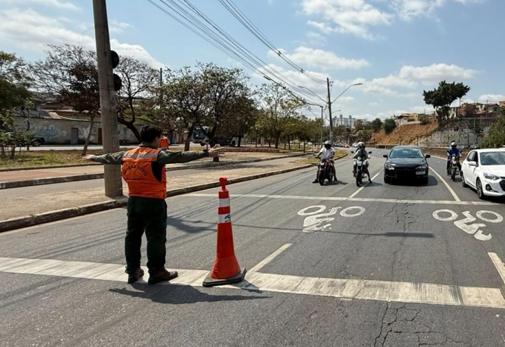 Defesa Civil realiza 6º simulado de fechamento de via na Avenida Bernardo Vasconcelos Imagem mostra um agente da Defesa Civil com colete laranja orientando o trânsito. Na foto há três motoqueiros parados e dois carros. Há árvores do lado esquerdo.