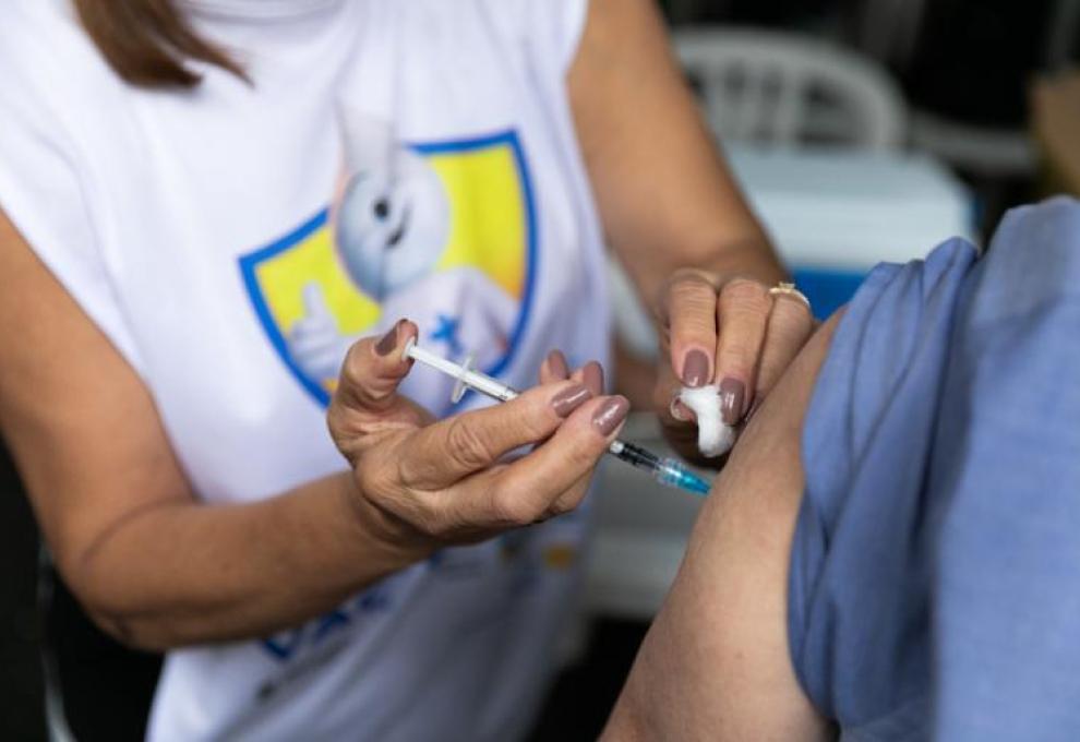 A foto mostra, em primeiro plano, uma profissional de saúde aplicando a vacina no braço de uma pessoa.