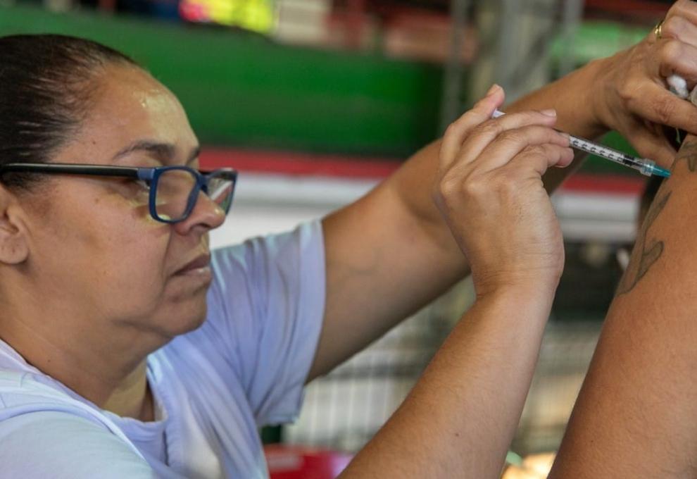 Mulher com blusa branca e óculos de aro preto aplica vacina no braço de uma pessoa