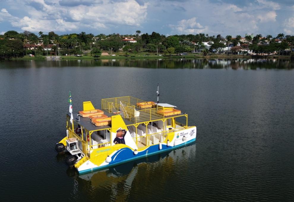 Barco turístico, nas cores branco, azul e amarelo, navegando na Lagoa da Pampulha, mostrando a orla ao fundo