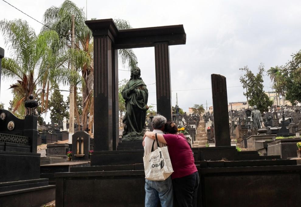 Em um cemitério, um casal abraçado aparece de costas diante de um imponente mausoléu de mármore preto, onde há uma estátua de bronze de uma figura em oração. A mulher veste camiseta vinho e o homem, camisa branca. Ao fundo, alinhamentos de túmulos sobem por uma encosta, acompanhados por palmeiras e pela vegetação do local. 