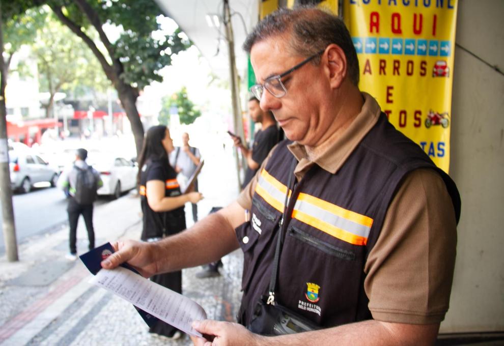 Fiscal de colete da Prefeitura de Belo Horizonte analisa um documento impresso na calçada durante ação de fiscalização de poluição visual na cidade.