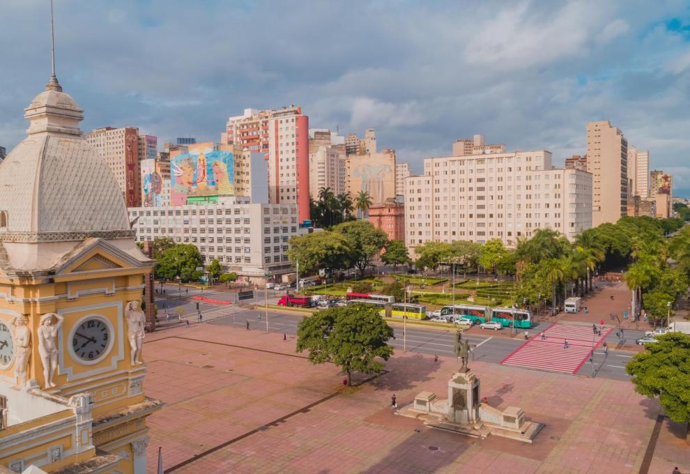 Vista panorâmica da Praça Rui Barbosa, no Centro de Belo Horizonte, com destaque para o centro da praça, de frente para a Avenida dos Andradas. Em primeiro plano, aparece a torre do antigo edifício da Estação Central, enquanto, ao fundo, prédios comerciais e residenciais compõem a paisagem urbana. A imagem também mostra áreas arborizadas, faixas de pedestres, veículos e ônibus em circulação, sob um céu parcialmente nublado.