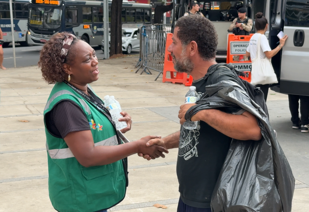 PBH reforça ações de conscientização e proteção diante de onda de calor na capital A foto mostra uma cena ao ar livre, em uma calçada movimentada de uma área urbana. No primeiro plano, duas pessoas conversam e apertam as mãos. À esquerda, uma mulher usa colete verde com faixas refletivas e segura uma garrafa de água. À direita, um homem veste camiseta preta e parece estar usando uma capa ou plástico escuro sobre os ombros; ele também segura uma garrafa de água. Ao fundo, há ônibus, veículos, agentes uniformizados, grades de isolamento e equipamentos de sinalização.
