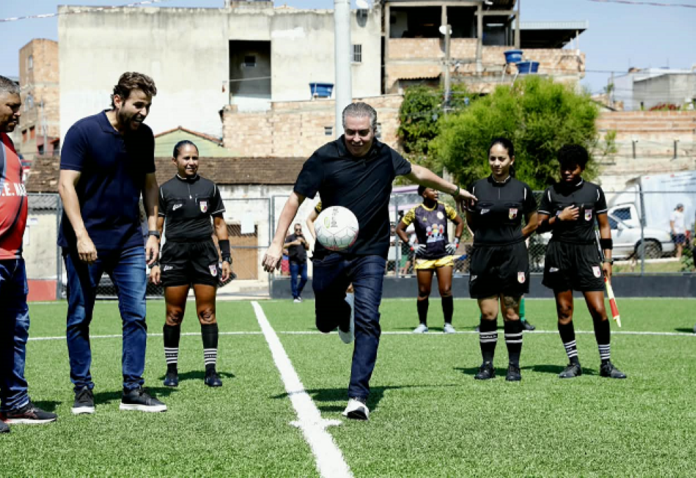 Homem branco,, prefeito Álvaro Damião, trajando calça jeans escura e camisa preta chuta uma bola de futebol branca em um campo de futebol. Ao lado dele um homem branco e três árbitras de futebol observam 