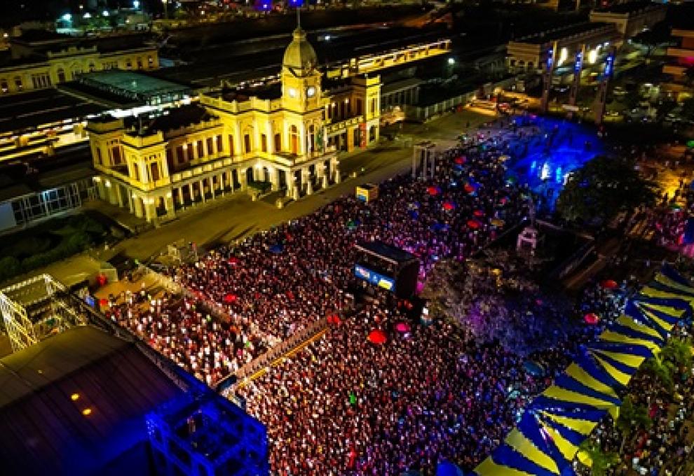 Início da celebração dos 129 anos de BH reúne mais de 20 mil pessoas em shows na Praça da Estação Imagem aérea noturna na Praça da Estação, em Belo Horizonte, tomada por um grande público durante a celebração pelo aniversário de 129 anos da capital mineira. Milhares de pessoas ocupam o espaço em frente ao Museu de Artes e Ofícios, iluminado por tons de amarelo. À direita, estruturas de palco, tendas azuis e amarelas e luzes coloridas compõem o cenário da festa.