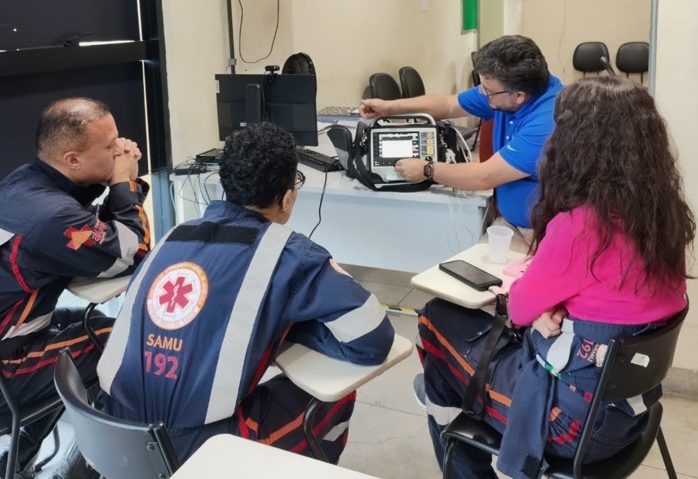 Tecnologia aproxima Samu e hospitais para aprimorar atendimento em casos de infarto Quarto profissionais do Samu, três homens e uma mulher, sentados em carteiras em círculo e um dos homens mostrando um aparelho tecnológico apoiado em cima de uma mesa.