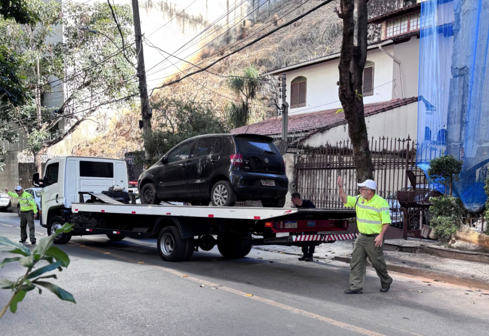 Um caminhão-guincho branco está parado em uma rua íngreme e gramada para rebocar um carro preto danificado em uma área residencial arborizada. Dois agentes da BHTrans coordenam a operação, com um deles sinalizando na frente do veículo e o outro posicionado na parte traseira.