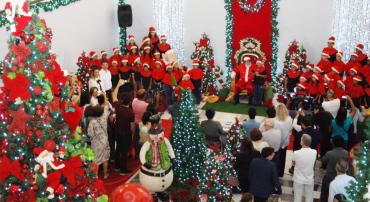 Mais de trinta estudantes municipais, vestidos com roupas natalinas, cantam nas laterais da cadeira de Papai Noel, na entrada da PBH. À frente, mais de vinte cidadãos assistem a apresentação. O local tem árvores e decoração natalina.
