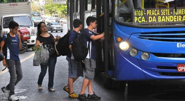 Dois estudantes entram em ônibus coletivo, durante o dia.