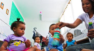 Educadora onfantil interage com quatro crianças com menos de um ano de idade com chocalho e brinquedos feitos como material reciclável. 