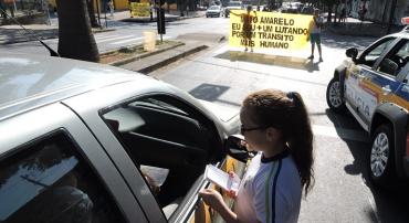 No primeiro plano, uma menina, aluna da rede municipal de ensino, entrega panfleto em carro. Ao fundo, dois alunos seguram uma faixa, em cima da faixa de pedestre, com os seguintes dizeres: "Maio amarelo: eu sou + um lutando por um trânsito mais humano".