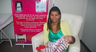Mãe sentada com criança recém nascida no colo. Ao fundo, cartaz do programa de aleitamento materno.