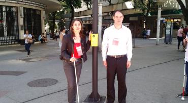 Dois Deficientes visuais aguardam ao lado do semáforo sonoro a hora de atravessar a rua.