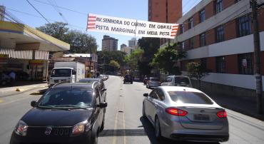 Carros transitam na Avenida Uruguai, próximo ao cruzamento com Avenida Nossa Senhora do Carmo; local tem faixa com os dizeres: Av. Nossa Senhora do Carmo em obras. Pista marginal em meia pista.