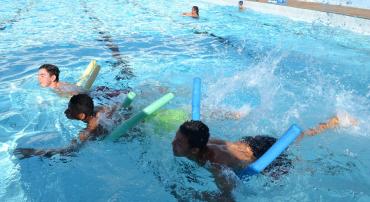 Três crianças fazem treinamento de natação apoiados bóia espaguete em em piscina, durante o dia. 