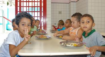 Cerca de oito crianças da Rede Municipal de Educação sentadas em uma mesa comendo merenda em pratos.