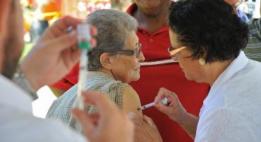 Senhora de idade recebe vacina contra a Influenza de técnica de saúde; mais a frente, detalhe das mãos de outro técnico, enchendo a seringa com vacina para outra aplicação.