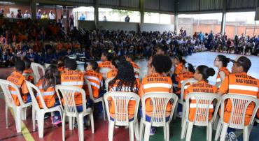 Cerca de vinte alunos de escola municipal usando colete laranja com os dizeres "Defesa Civil nas escolas" e se apresentam para outras centenas de alunos