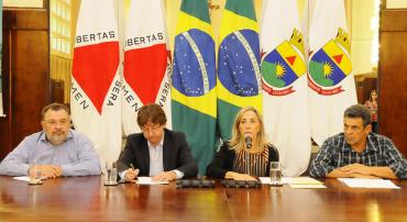 Foto mostra a secretária de Política Urbana, Maria Caldas, falando ao microfone. Ao fundo, as bandeiras de MG, BH e do Brasil.