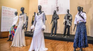 Nove manequins com diferentes vestidos. O vestido em destaque é branco e tem plumas nas mangas.