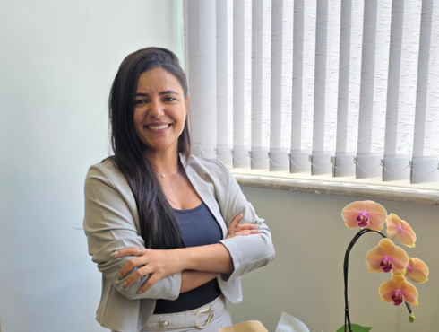 Mulher sorridente, vestindo blazer bege sobre blusa azul, com os braços cruzados, em um ambiente de escritório com persianas ao fundo e uma orquídea sobre a mesa.