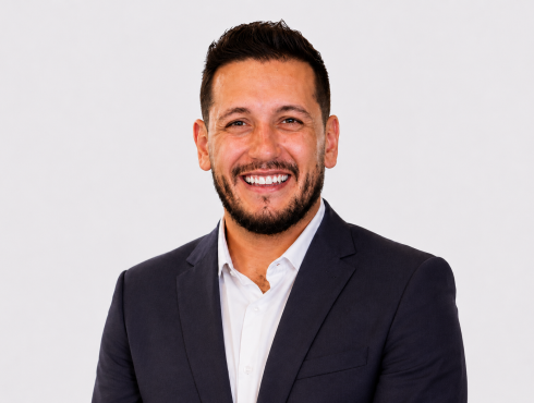 Homem de cabelos curtos e escuros, barba bem aparada, pele clara, olhos escuros e sorriso amplo. Veste camisa branca e blazer escuro, com aparência profissional e descontraída.