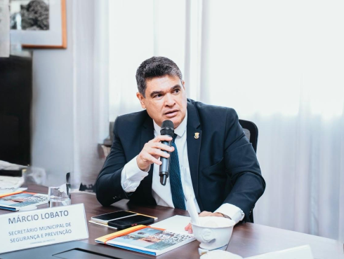 Homem sentado à mesa, usando terno escuro, camisa branca e gravata azul, segurando um microfone enquanto fala. Ao fundo, há uma janela com cortinas claras e, sobre a mesa, estão documentos, celulares, um copo e uma pequena placa com a identificação “Márcio Lobato — Secretário Municipal de Segurança e Prevenção”