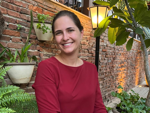 Mulher adulta, de cabelos escuros presos, pele clara e olhos castanhos. Apresenta um sorriso simpático e veste uma blusa de manga longa na cor vinho. Está em um ambiente externo com plantas e parede de tijolos aparentes.