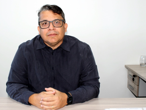 Ricardo Luiz de Lima - Diretor de Edificações e Espaços Urbanos da Sudecap Homem de meia-idade, de pele clara, com cabelos curtos escuros e fios grisalhos nas laterais. Usa óculos de armação preta, camisa social de manga longa em tom escuro, relógio inteligente no pulso esquerdo e aliança. Está sentado à mesa com as mãos entrelaçadas, olhando diretamente para a câmera, com expressão neutra e postura formal.