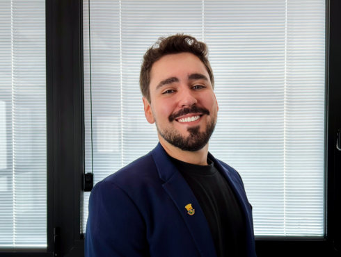 Luís Castilho Homem de aparência jovem, com cabelos curtos castanhos, barba e bigode. Está vestido com blazer azul-marinho sobre camiseta preta, usando um broche na lapela, em um ambiente corporativo moderno e bem iluminado. A imagem passa uma impressão de credibilidade, acessibilidade e profissionalismo.