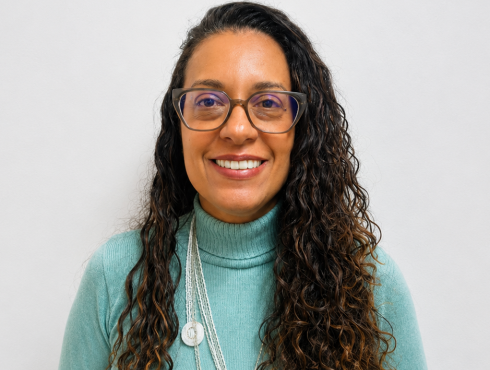 Alessandra Duarte Rodrigues Pereira Mulher de cabelos longos, escuros e cacheados, usando óculos de armação escura. Está sorrindo, veste uma blusa de gola alta verde-claro e um colar longo branco, em frente a um fundo claro e neutro.