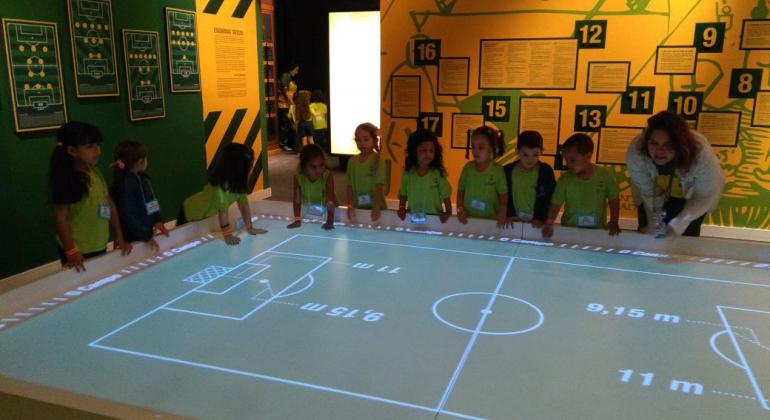 Educando a Cidade para Educar / Foto: Magi Mappa/PBH Nove crianças, alunos da educação infantil, e uma adulta, em volta de uma tela com um campo de futebol