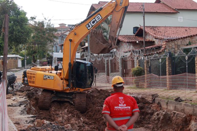 Homem de uniforme vermelho parado de frente para uma trator retirando terra 