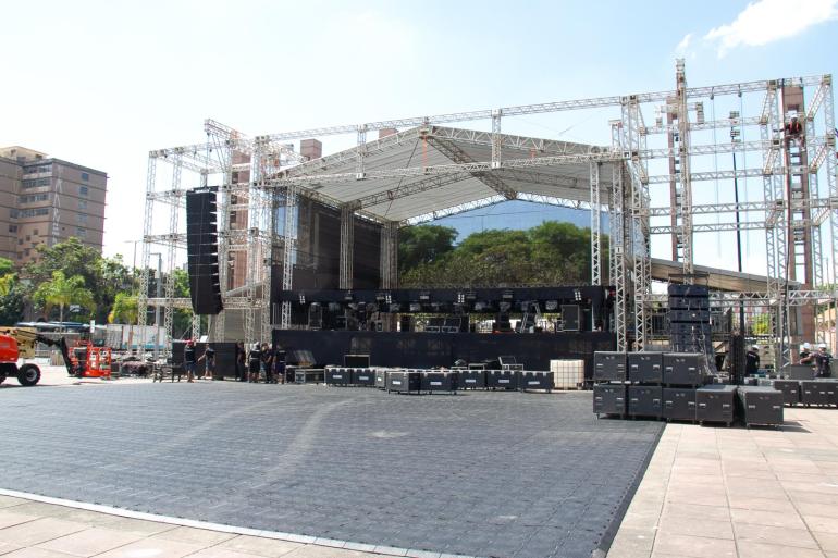 Operação de trânsito para a festa aniversário de Belo Horizonte, na Praça da Estação A foto mostra a montagem de um grande palco para um evento ao ar livre, em uma área ampla e pavimentada. A estrutura é composta por treliças metálicas que sustentam a cobertura, os equipamentos de iluminação e os sistemas de som. Nas laterais do palco, há grandes conjuntos de caixas de som, enquanto diversos equipamentos técnicos estão distribuídos na parte frontal e interna da estrutura. No chão, em frente ao palco, há uma extensa área coberta por placas escuras, provavelmente destinada ao público.