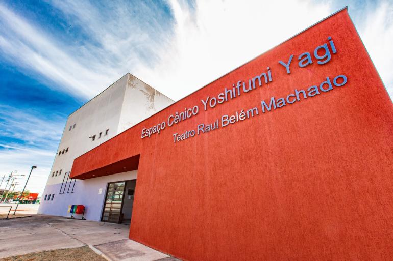 Fachada do teatro vista de baixo para cima, com parede de textura avermelhada e a inscrição em "Espaço Cênico Yoshifumi Yagi" em destaque no topo. Abaixo outro letreiro "Teatro Raul Belém Machado"
