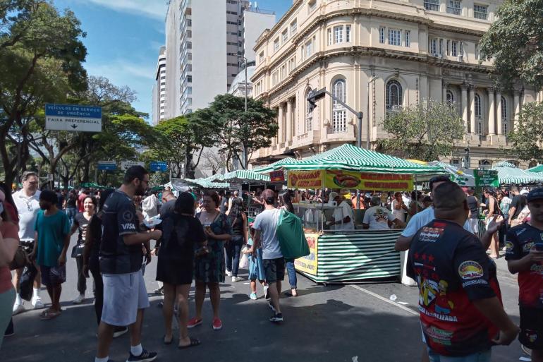 Diversas barracas de alimentação cobertas por lonas listradas em verde e branco. Ao centro e ao fundo, várias pessoas circulam, compram alimentos e conversam 